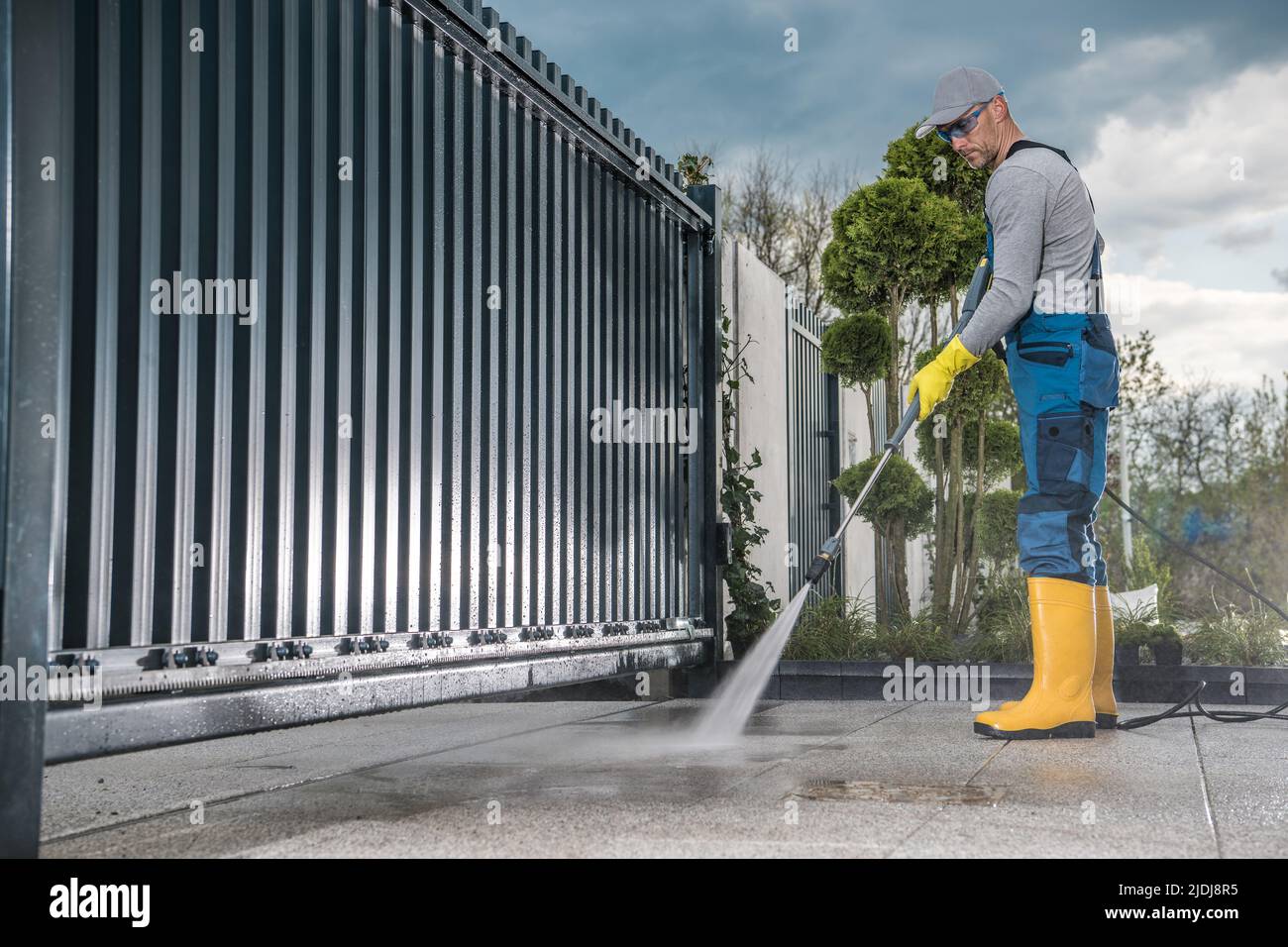 Travailleur caucasien dans son travail porter des bottes en caoutchouc jaune nettoyer l'allée en béton devant la porte d'entrée à l'aide d'un lavage à pression professionnel Banque D'Images