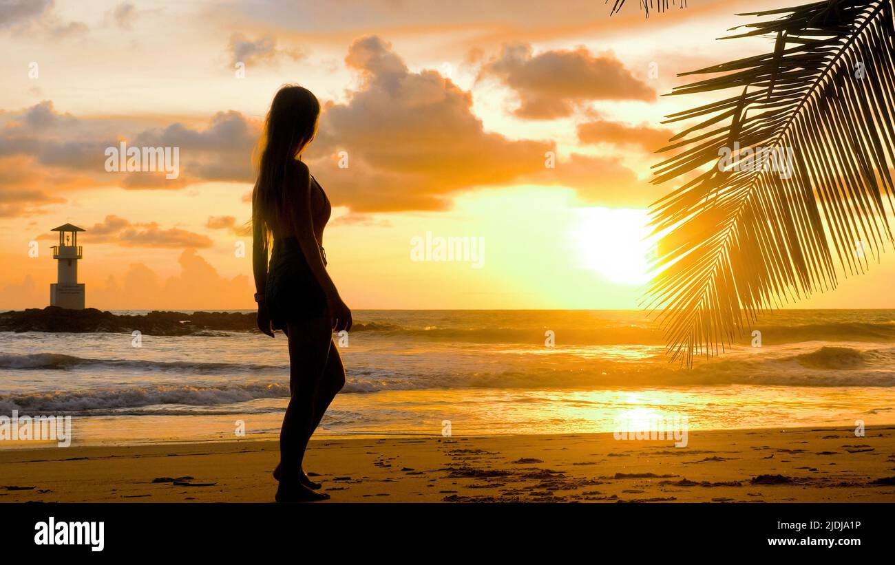 Une femme touriste marchant pieds nus le long des vagues d'eau de mer sur une plage de sable. Une jeune femme marche au bord de la mer dans des lumières dorées romantiques au coucher du soleil. Bonne fille s'amuser Banque D'Images