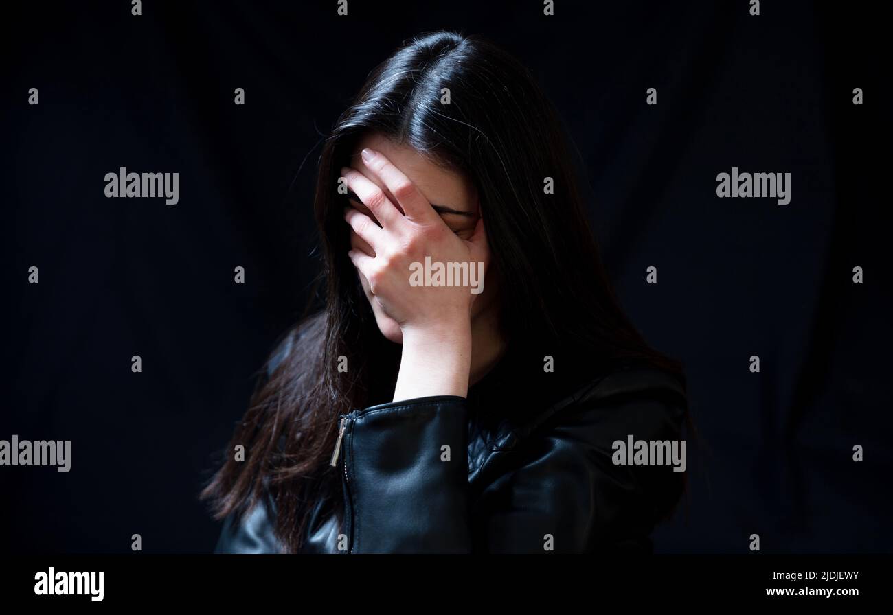 Jeune femme en cuir noir blouson couvrant les yeux, honte et regret émotion dans une pièce sombre. Concept de peur et de désespoir Banque D'Images