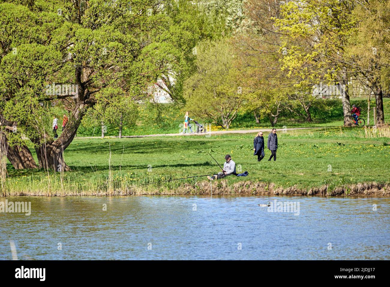 Riga, Lettonie - 8 mai 2022: Un pêcheur est assis avec une canne à pêche sur la rive d'un lac dans un parc public. Herbe verte autour. Les gens marchent à distance. Banque D'Images