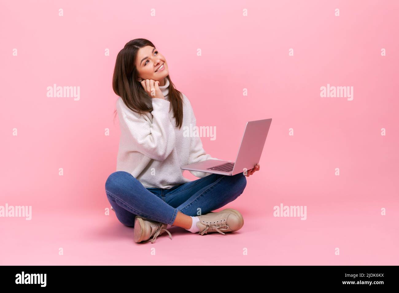 Portrait d'une femme avec une expression faciale rêveuse, assise avec des jambes croisées sur le sol, tenant un ordinateur portable, portant un pull blanc de style décontracté. Studio d'intérieur isolé sur fond rose. Banque D'Images