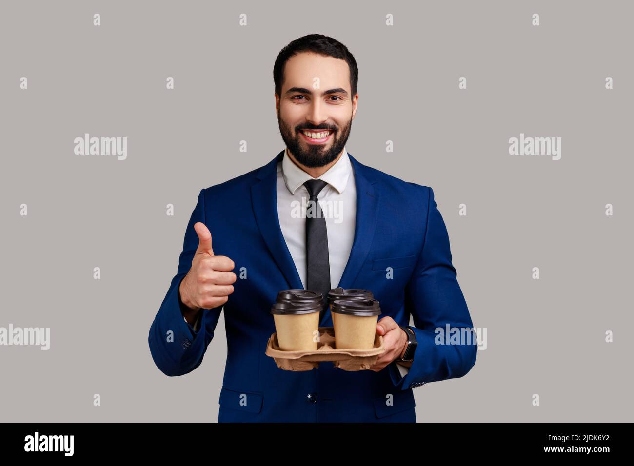Homme optimiste montrant le pouce vers le haut, tenant deux gobelet en papier avec une boisson, veut boire du café pendant la pause avec un ami, portant le costume de style officiel. Prise de vue en studio isolée sur fond gris. Banque D'Images