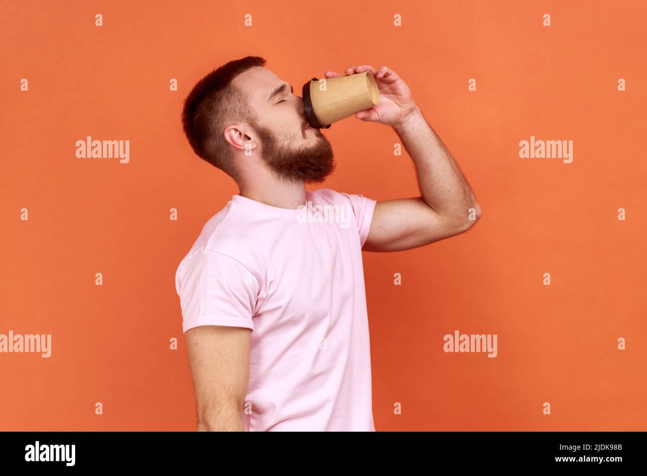 Vue latérale portrait de l'homme endormi et fatigué barbu un café de tasse jetable, besoin d'énergie le matin, portant un T-shirt rose. Studio d'intérieur isolé sur fond orange. Banque D'Images