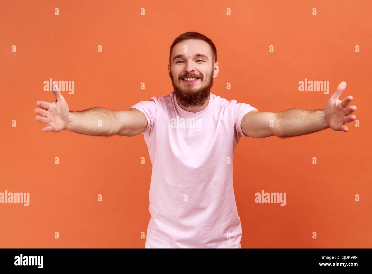 Entrez dans mes bras. Portrait d'un homme barbu heureux et positif qui se fixe à l'appareil photo, qui s'étire les bras pour vous embrasser, en portant un T-shirt rose. Studio d'intérieur isolé sur fond orange. Banque D'Images