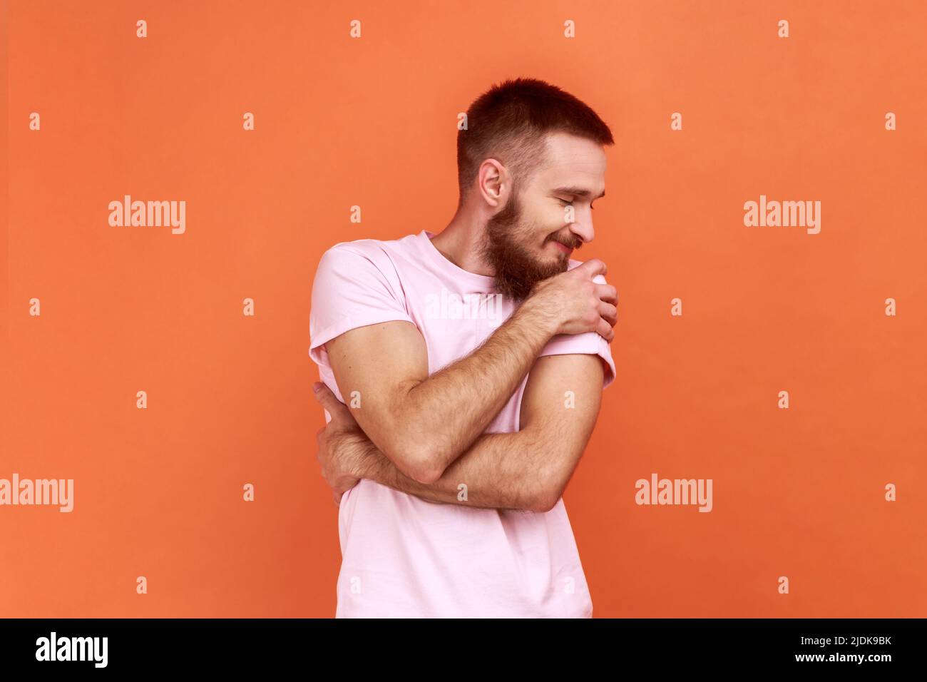 Portrait de l'homme à barbe heureux s'embrassant et souriant avec l'expression du grand ego, du plaisir et de l'estime de soi, portant un T-shirt rose. Studio d'intérieur isolé sur fond orange. Banque D'Images
