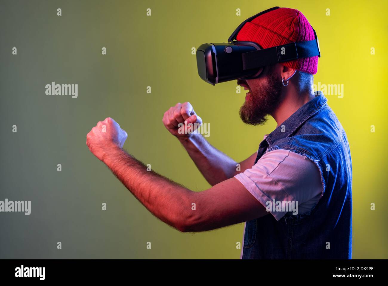 Vue latérale de l'homme hipster dans des lunettes de réalité virtuelle à la tête de jeu de combat, tenant les poings serrés prêts à la boxe. Studio d'intérieur isolé sur fond de néon coloré. Banque D'Images