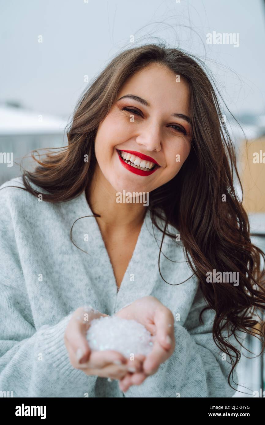 Jeune heureuse souriante merveilleuse femme avec long cheveux ondulés foncés maquillage porter un gilet gris, tenant des flocons de neige dans les paumes. Banque D'Images