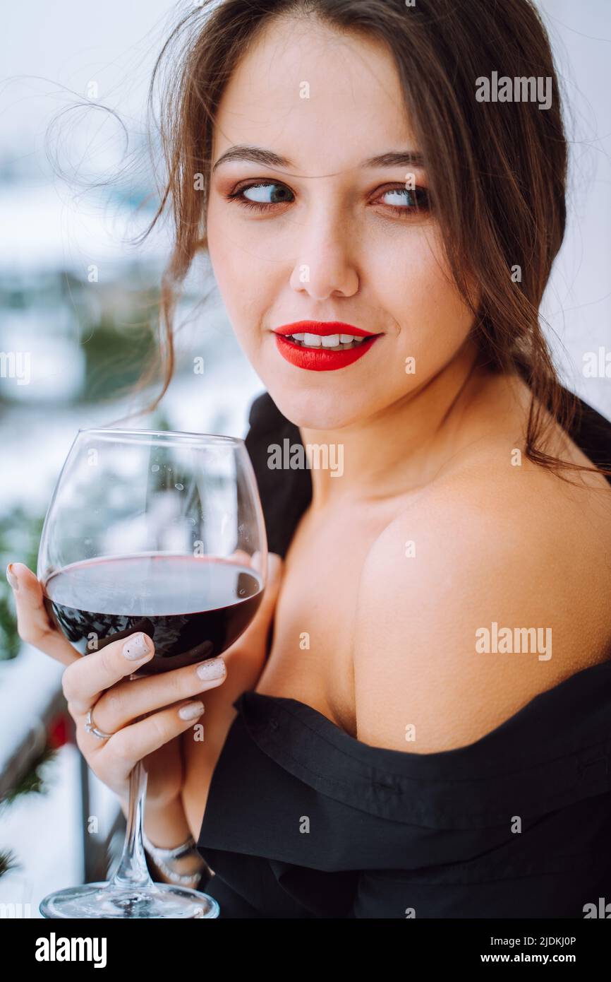 Jeune femme souriante porte une chemise noire avec une épaule nue debout sur le balcon, tenant le verre avec le vin rouge, regardant de côté. Banque D'Images