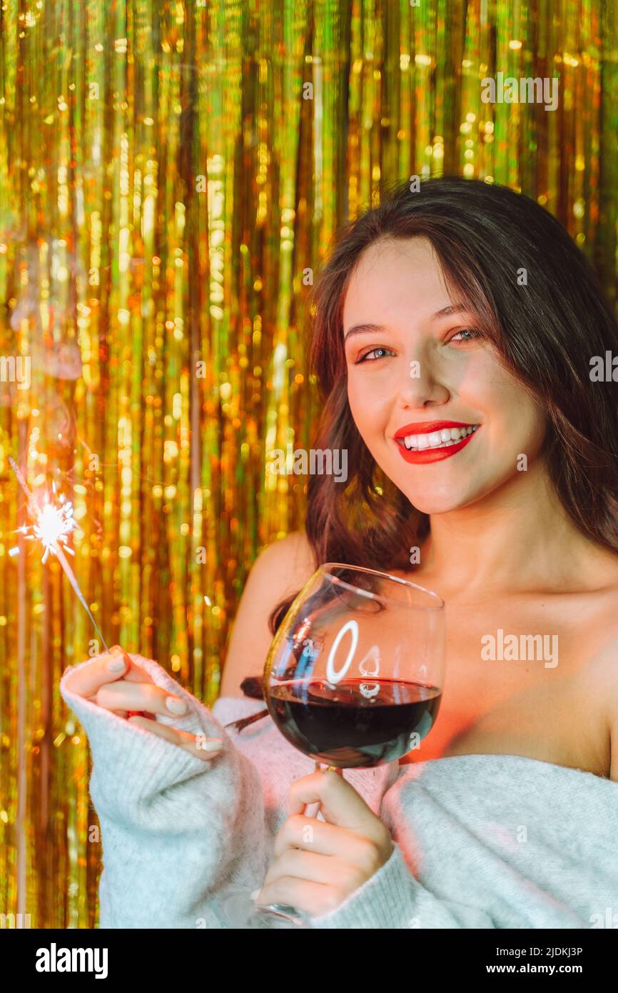 La jeune femme porte un gilet gris à épaules nues, avec un verre de vin rouge, un sparkler sur fond scintillant doré. Banque D'Images