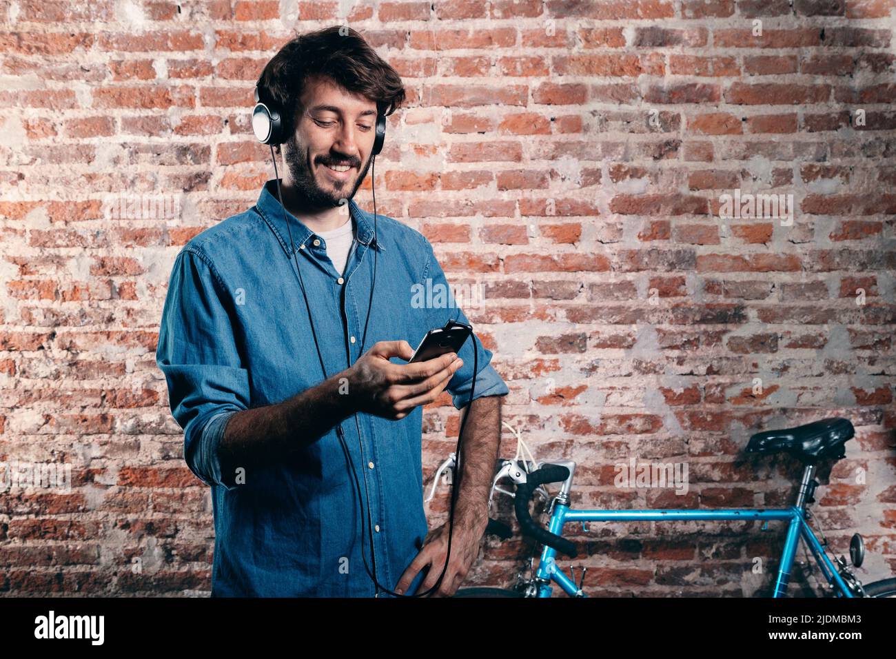Jeune millénaire Écoutez de la musique avec un téléphone portable et un casque. En arrière-plan, un vélo bleu appuyé contre le mur de briques. Banque D'Images