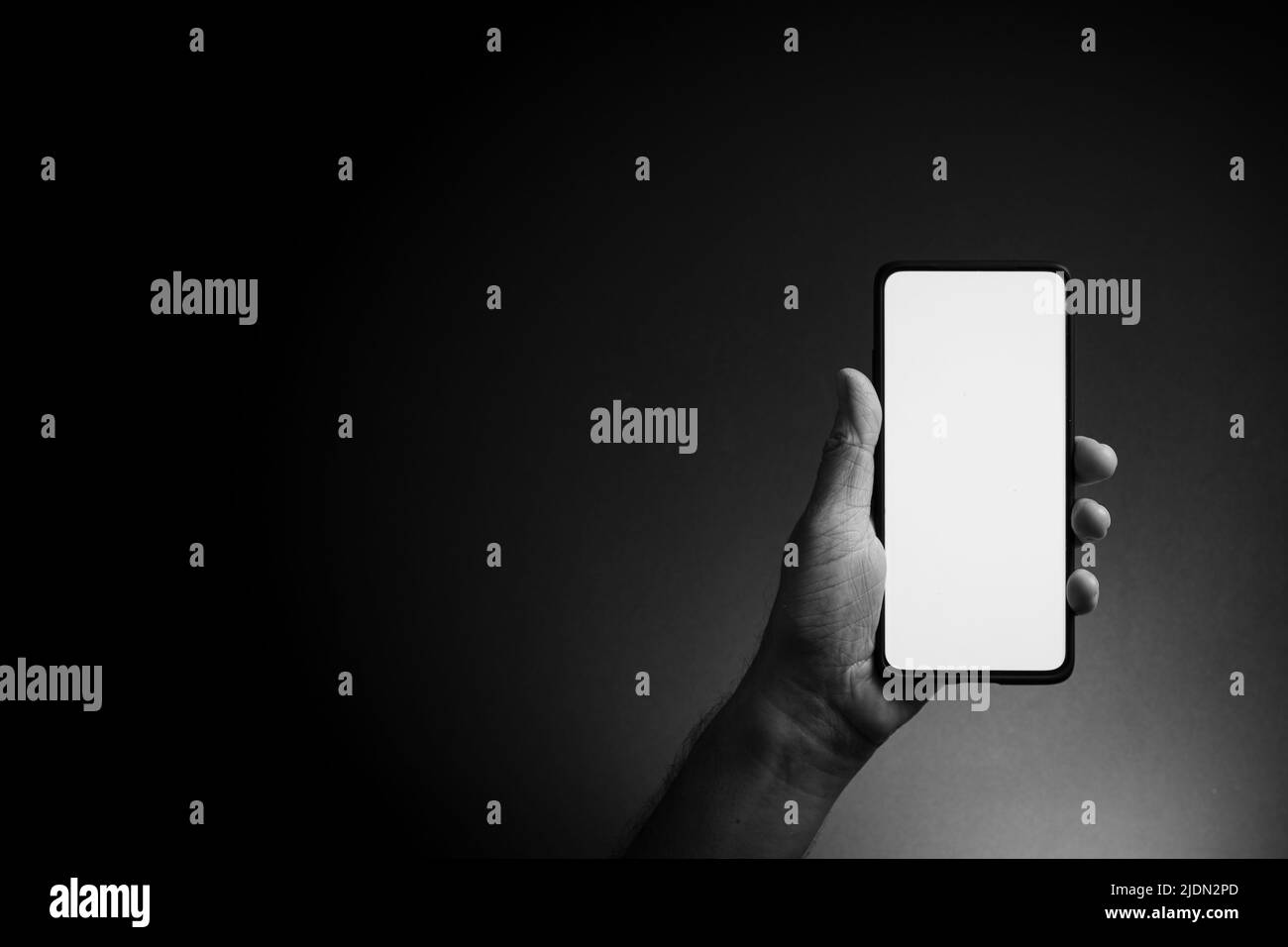 Image en noir et blanc de la main de l'homme tenant le smartphone noir verticalement avec écran blanc vierge isolé sur fond sombre avec un éclairage spectaculaire et Banque D'Images