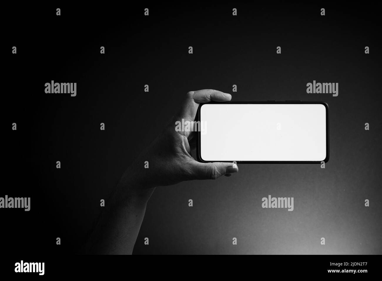 Image en noir et blanc de la main de l'homme tenant le smartphone noir horizontalement avec écran blanc vierge isolé sur fond sombre avec un éclairage spectaculaire a Banque D'Images