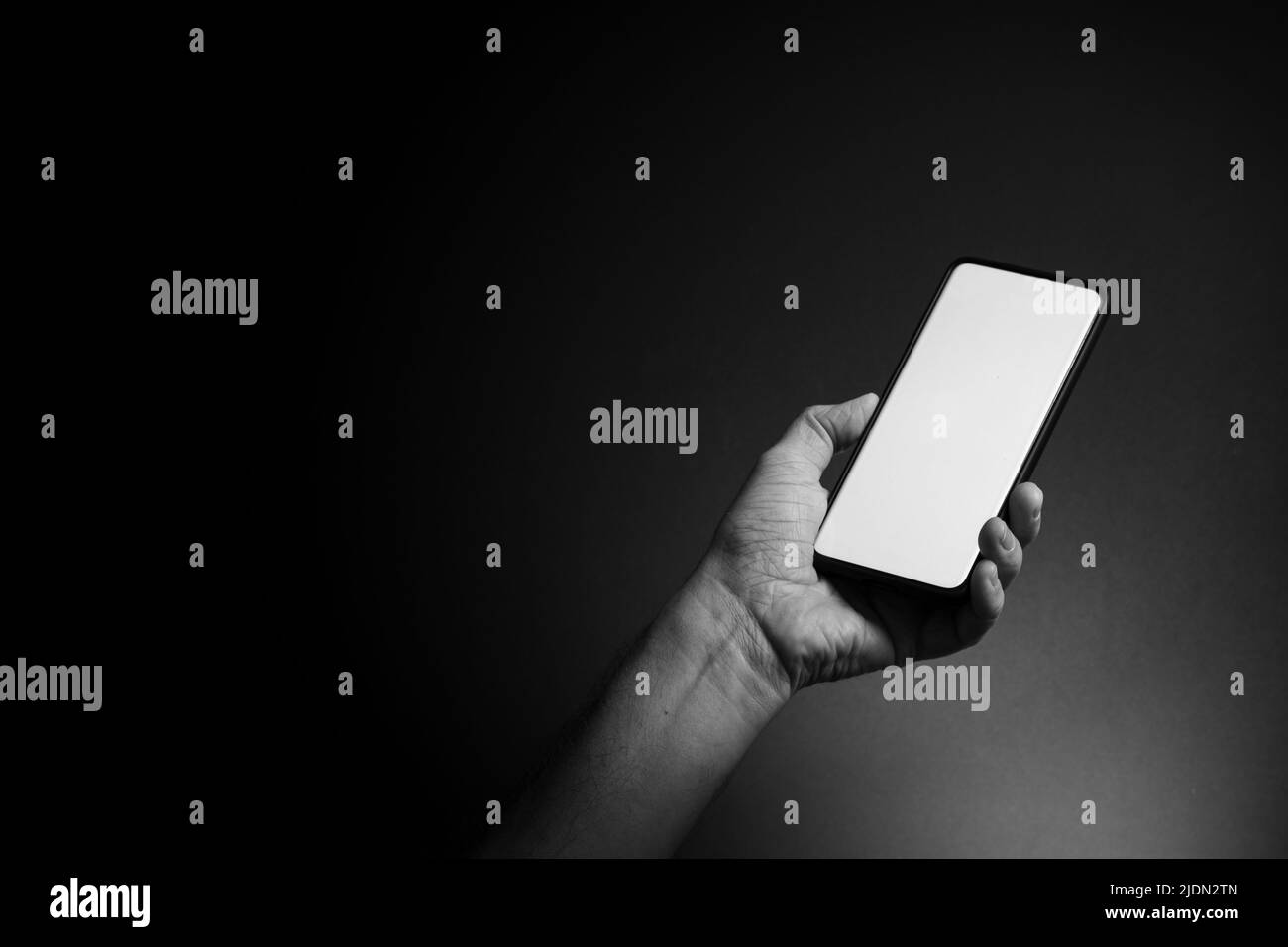 Image en noir et blanc de la main de l'homme tenant un smartphone noir avec écran blanc vierge isolé sur fond sombre avec un éclairage spectaculaire et un espace de copie Banque D'Images
