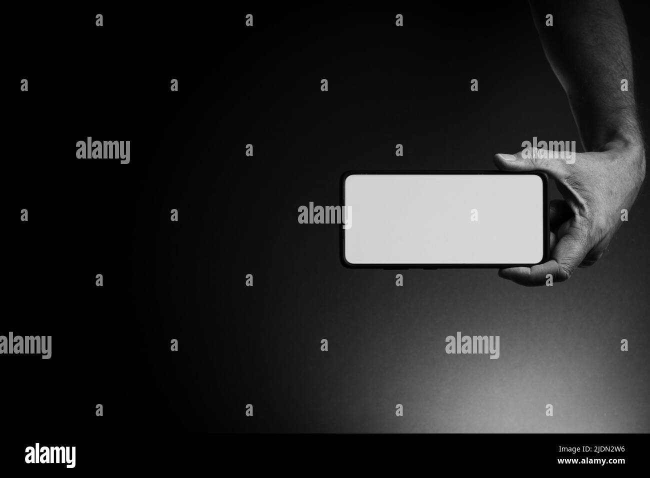 Image en noir et blanc de la main de l'homme tenant le smartphone noir horizontalement avec écran blanc vierge isolé sur fond sombre avec un éclairage spectaculaire a Banque D'Images