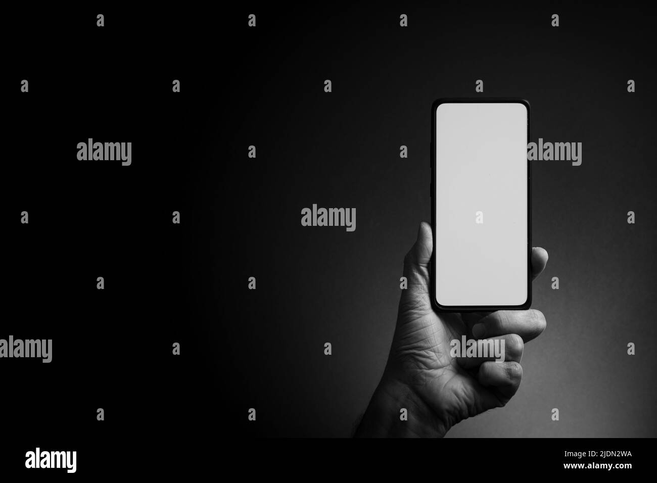 Image en noir et blanc de la main de l'homme tenant le smartphone noir verticalement avec écran blanc vierge isolé sur fond sombre avec un éclairage spectaculaire et Banque D'Images