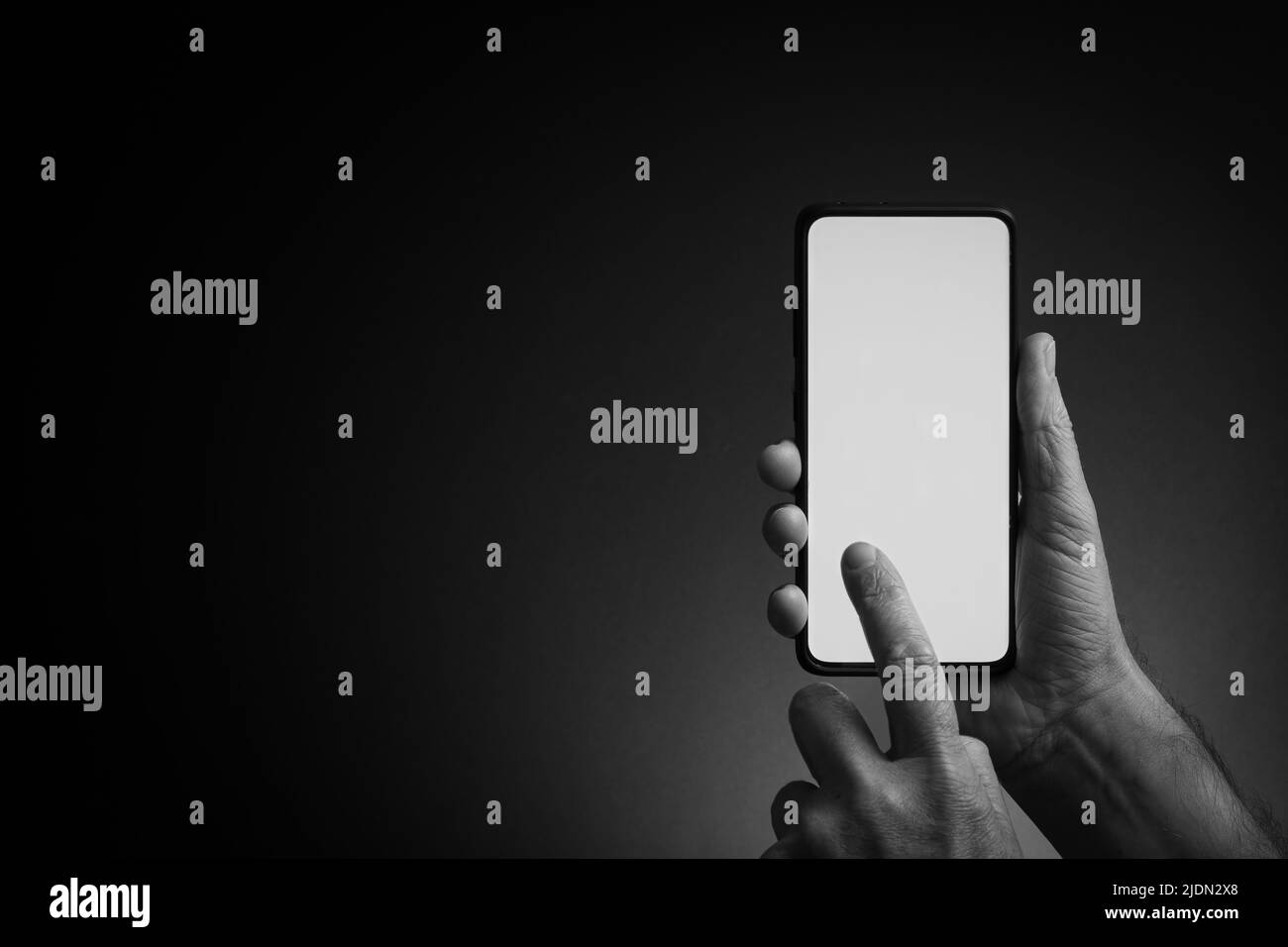 Image en noir et blanc de la main de l'homme tenant le smartphone noir et utilisant un écran blanc vierge avec le doigt isolé sur un arrière-plan sombre avec un éclairage spectaculaire Banque D'Images