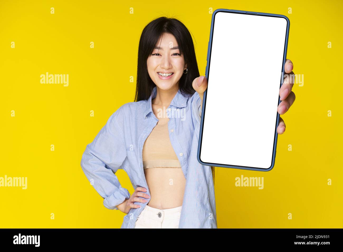 Fille asiatique tenant un smartphone montrant un écran blanc, et sourire excité sur l'appareil photo portant une chemise bleue isolée sur fond jaune. Offre exceptionnelle sur les applications mobiles. Positionnement du produit. Banque D'Images