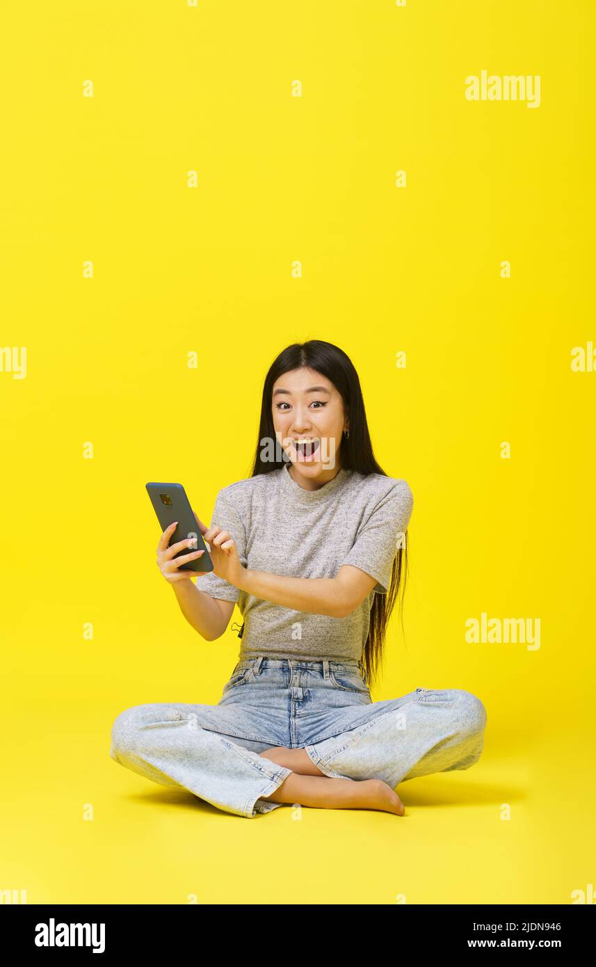 Une jeune fille asiatique excitée assise sur le sol tenant un smartphone texting ou magasiner en ligne ou gagner des jeux isolés sur fond jaune. Positionnement du produit. Publicité sur les applications mobiles. Copier l'espace. Banque D'Images