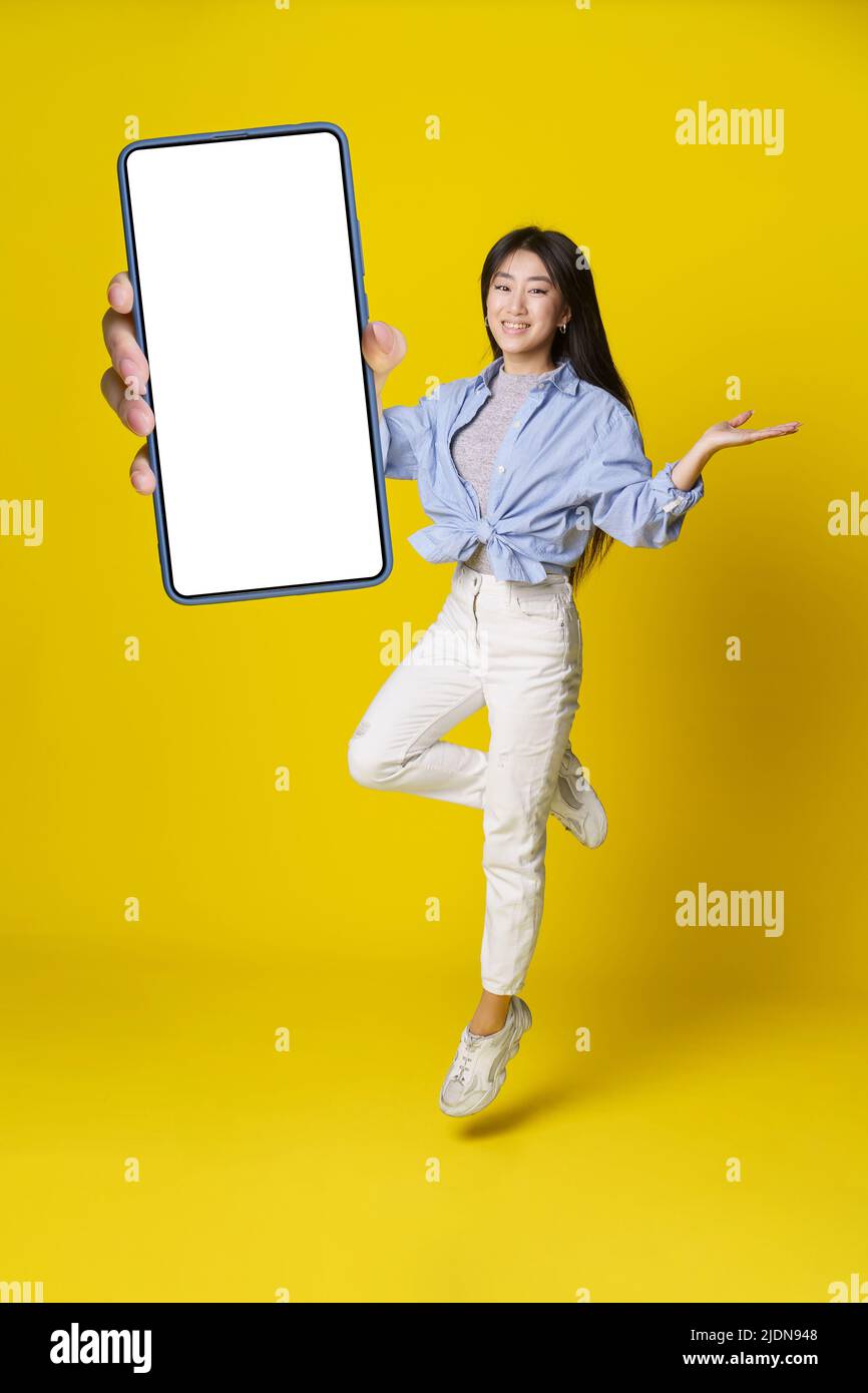 Fille asiatique en jump tenant un smartphone avec écran noir à la main dans une tenue décontractée, publicité d'application mobile isolée sur fond jaune. Portrait complet d'une fille asiatique joyeuse. Positionnement du produit. Banque D'Images