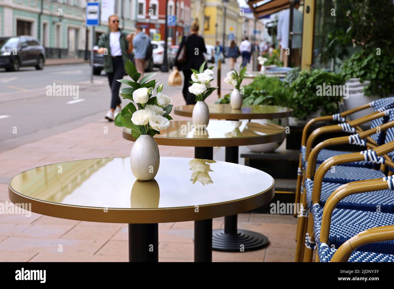 Street café dans la ville d'été avec des tables humides vides à l'extérieur sur le fond des gens de marche. Vases de fleurs roses sur des tables rondes et des chaises après la pluie Banque D'Images