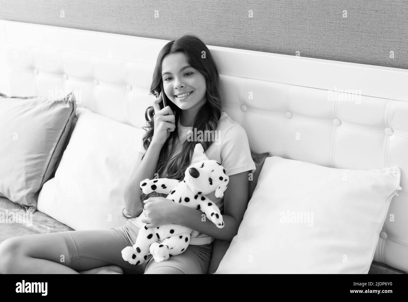 Un enfant heureux parle sur un téléphone portable assis avec un chiot jouet dans le lit, conversation téléphonique Banque D'Images