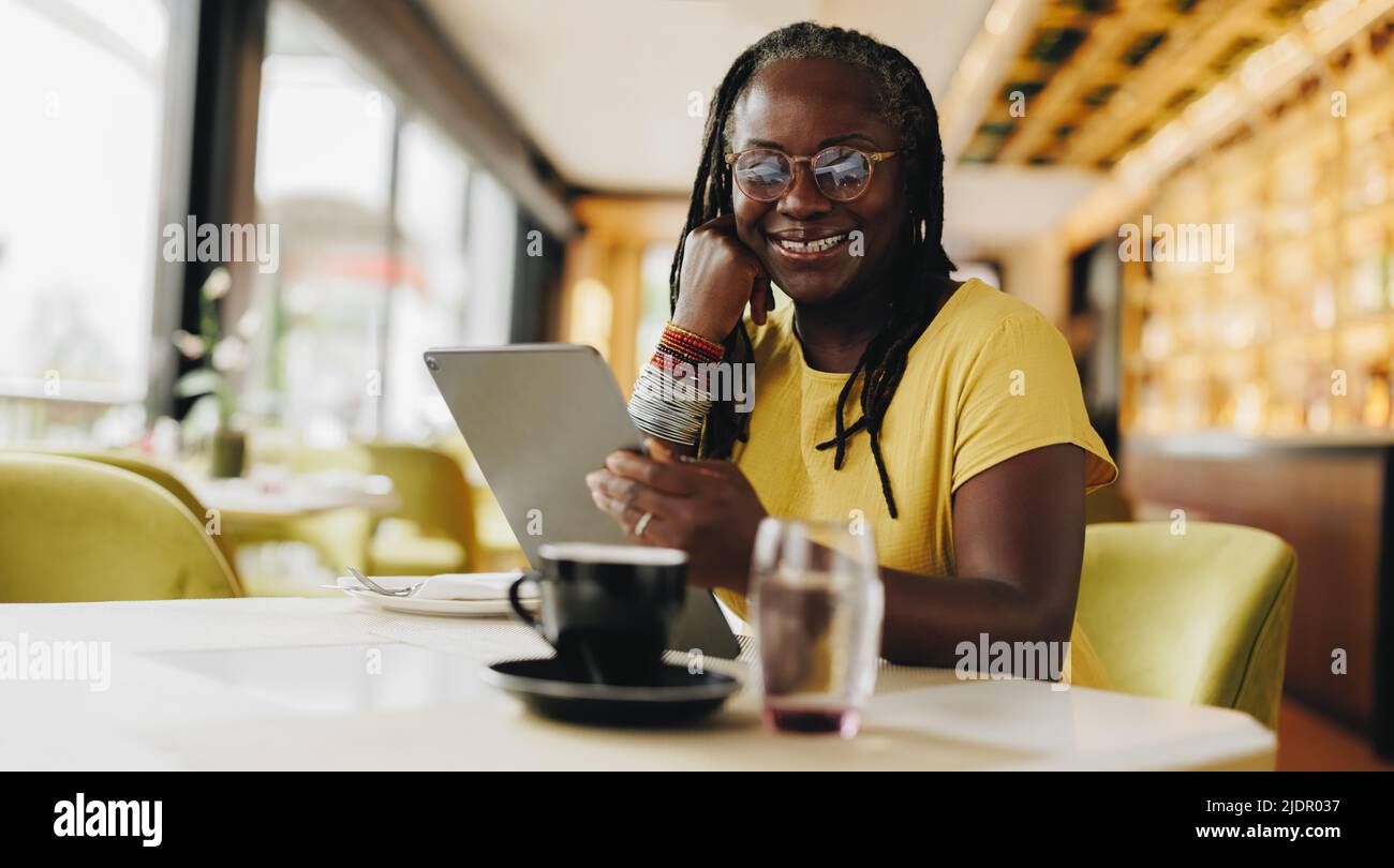 Bonne femme d'affaires souriant à la caméra tout en tenant une tablette numérique dans un café. Femme indépendante senior faisant un certain travail indépendant en ligne dans un c Banque D'Images
