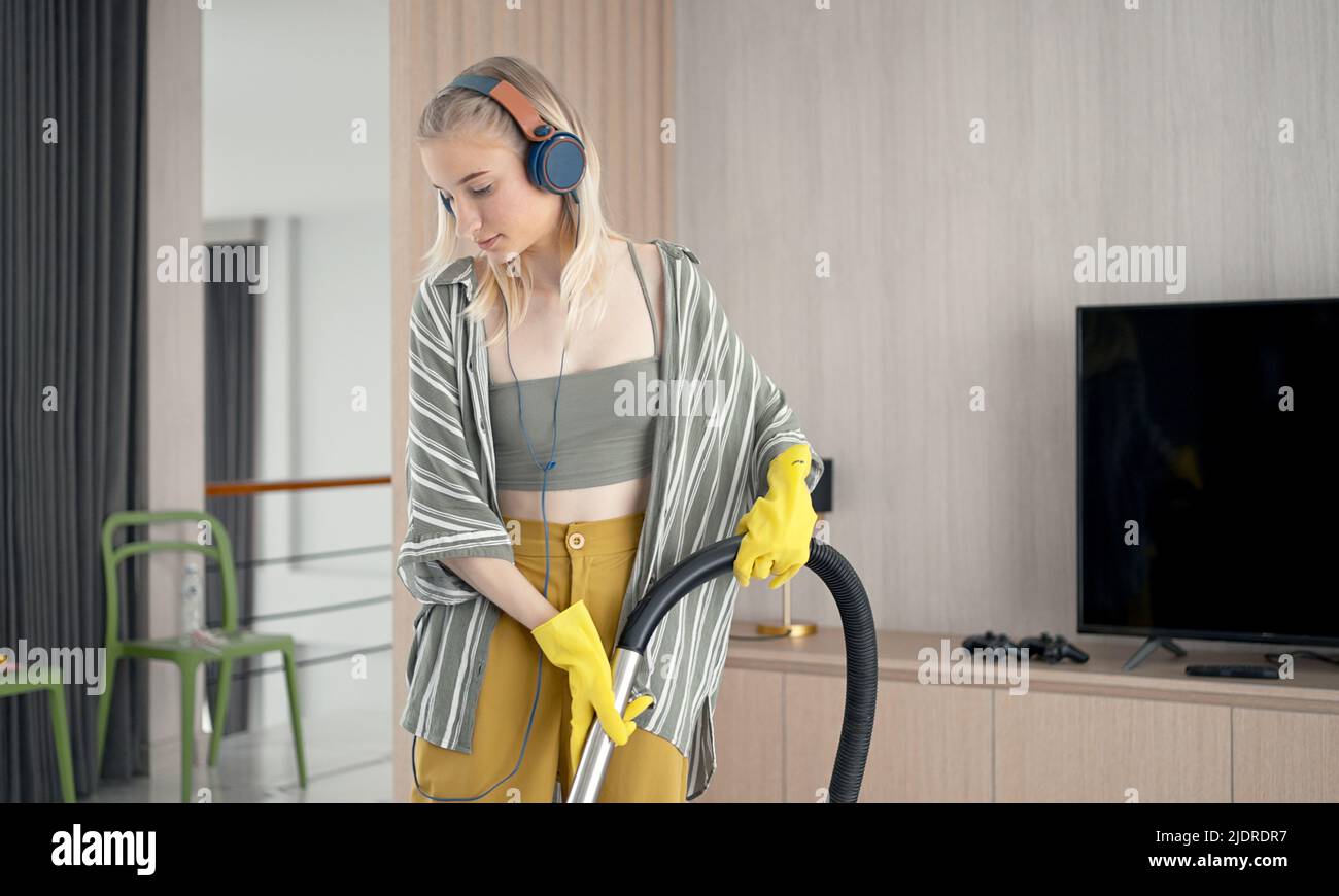 Une jeune fille se moque de nettoyer le sol avec un aspirateur. Bonne femme qui fait du ménage à la maison Profitez de la musique avec des écouteurs. Banque D'Images