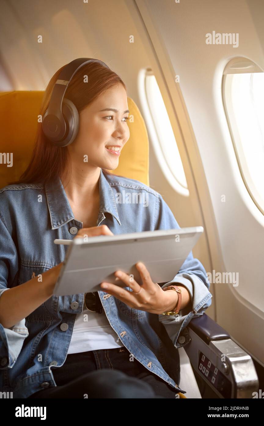 Charmante femme asiatique du millénaire à la place de sa fenêtre avec son casque sans fil utilisant une tablette portable et regardant par la fenêtre de l'avion. Banque D'Images