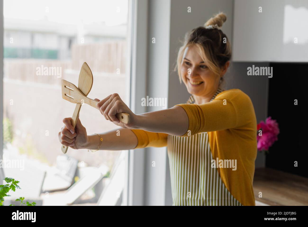 Drôle, inspiré, souriante femme blonde joyeuse de cuisine, croisant la spatule de cuisine en bois rigide armé. Cours de formation de cuisinier Banque D'Images