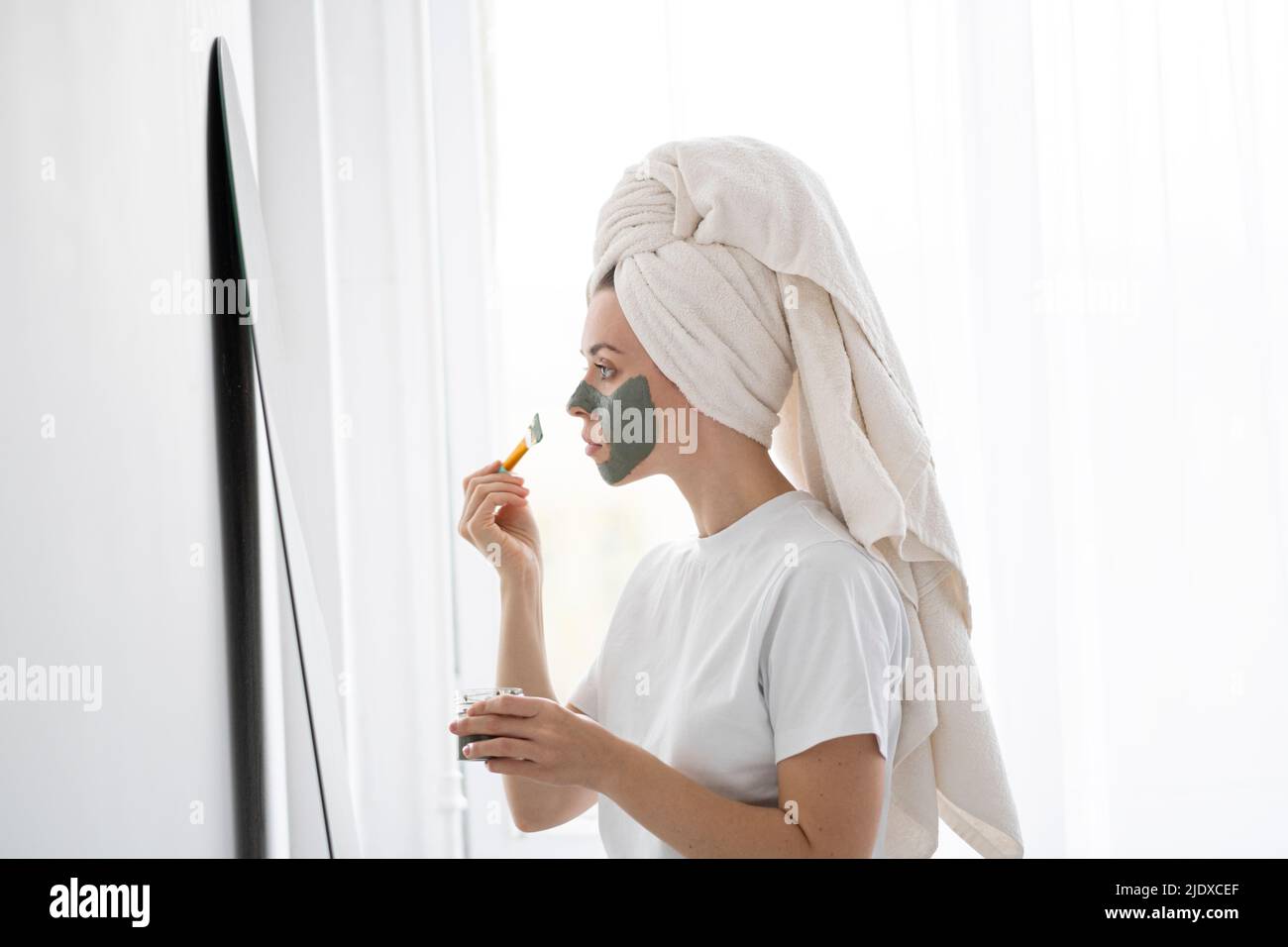 Jeune femme appliquant le masque facial devant le miroir à la maison Banque D'Images