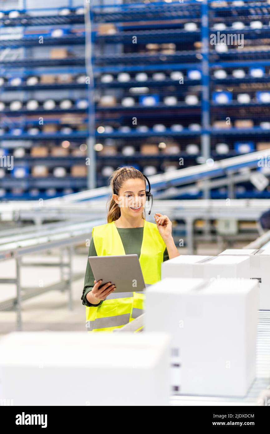 Jeune travailleur souriant avec un Tablet PC qui parle à travers un micro-casque situé près du tapis roulant dans l'entrepôt Banque D'Images