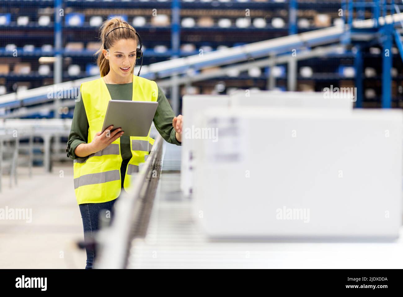 Jeune employé avec un PC tablette examinant des boîtes sur le tapis transporteur dans l'entrepôt Banque D'Images
