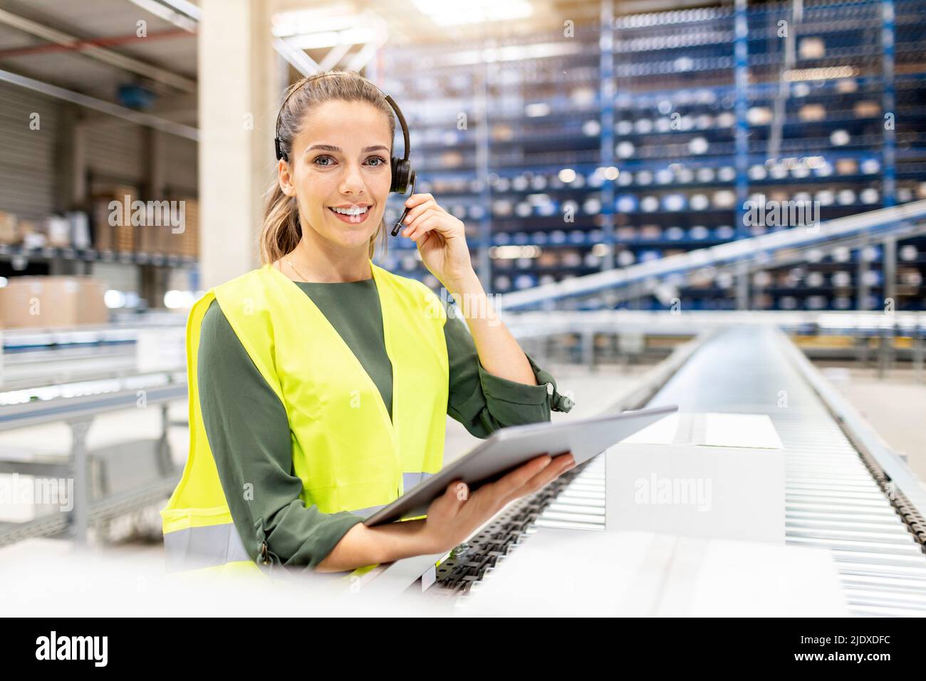 Travailleur souriant portant un casque debout avec un Tablet PC près du tapis roulant dans l'entrepôt Banque D'Images