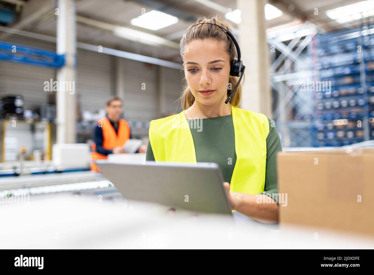 Jeune travailleur portant un micro-casque/une oreillette utilisant un PC tablette dans une boîte en carton dans l'entrepôt Banque D'Images