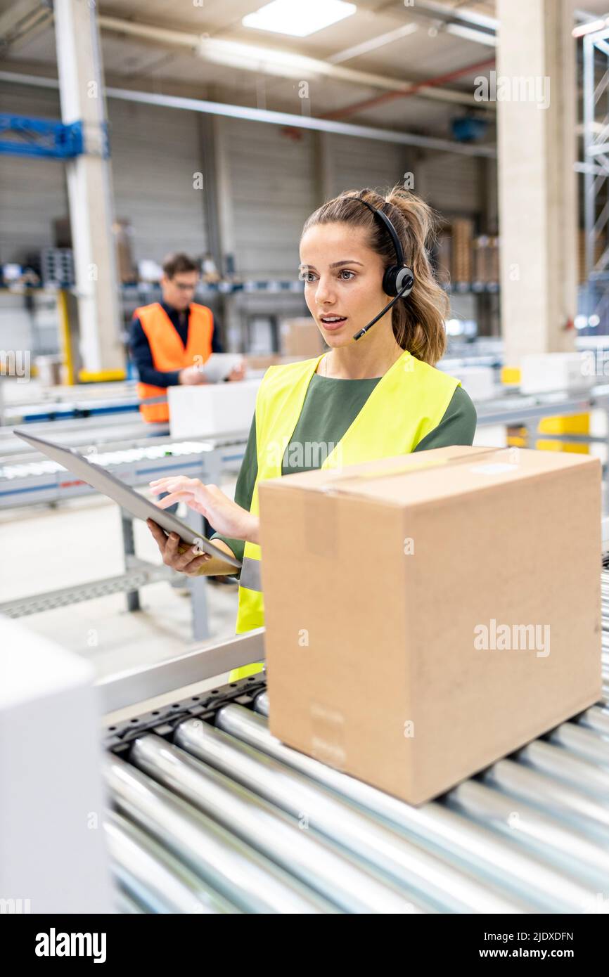 Jeune travailleur avec un PC tablette examinant des boîtes en carton sur le tapis transporteur dans l'entrepôt Banque D'Images