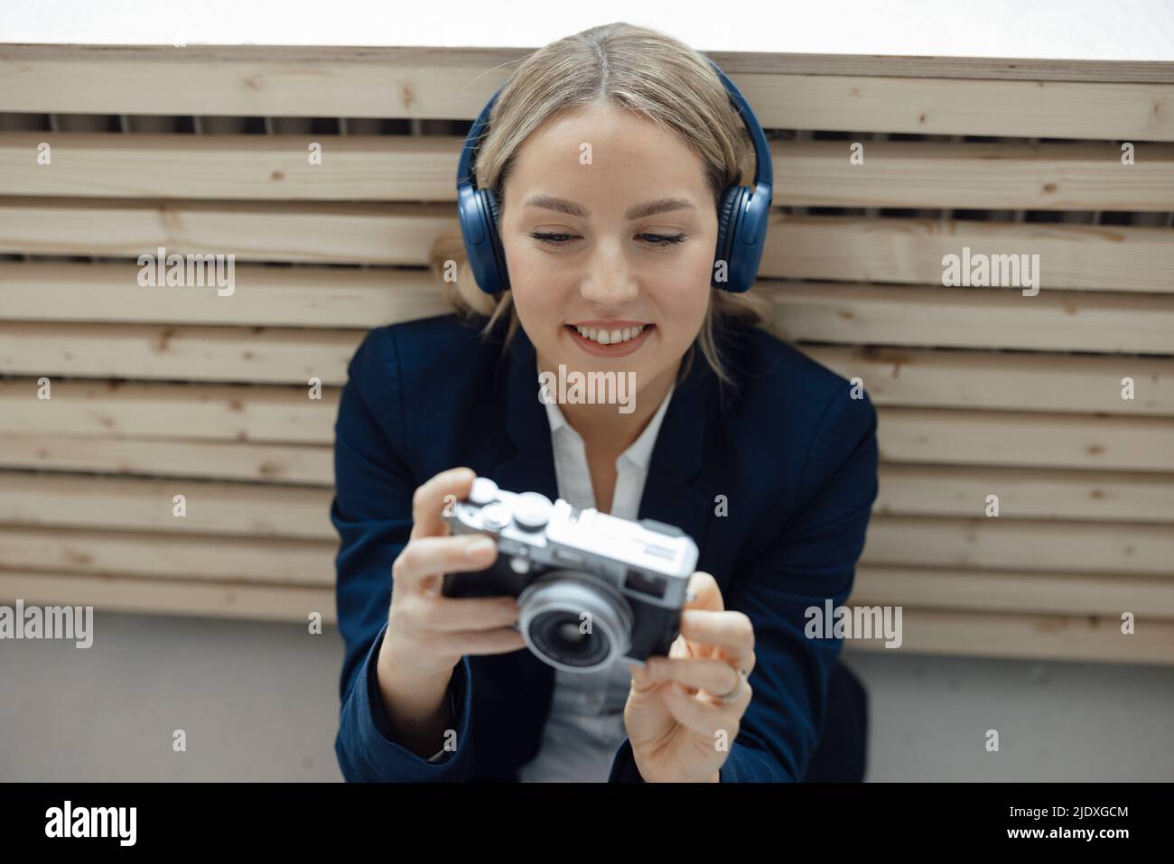 Femme d'affaires souriante tenant un appareil photo pour écouter de la musique à l'aide d'un casque sans fil au bureau Banque D'Images
