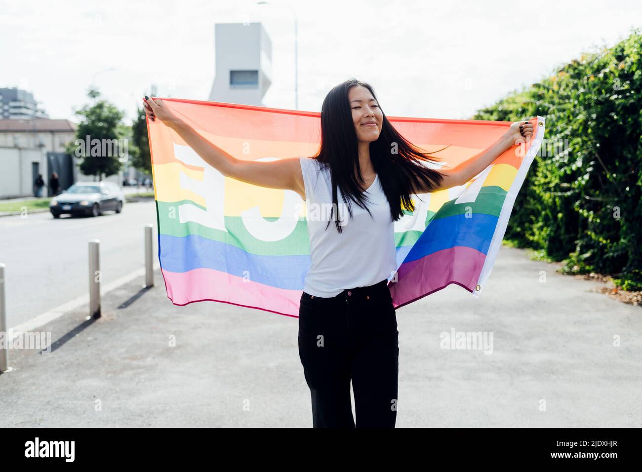 Femme souriante avec les yeux fermés tenant le drapeau arc-en-ciel marchant le jour ensoleillé Banque D'Images