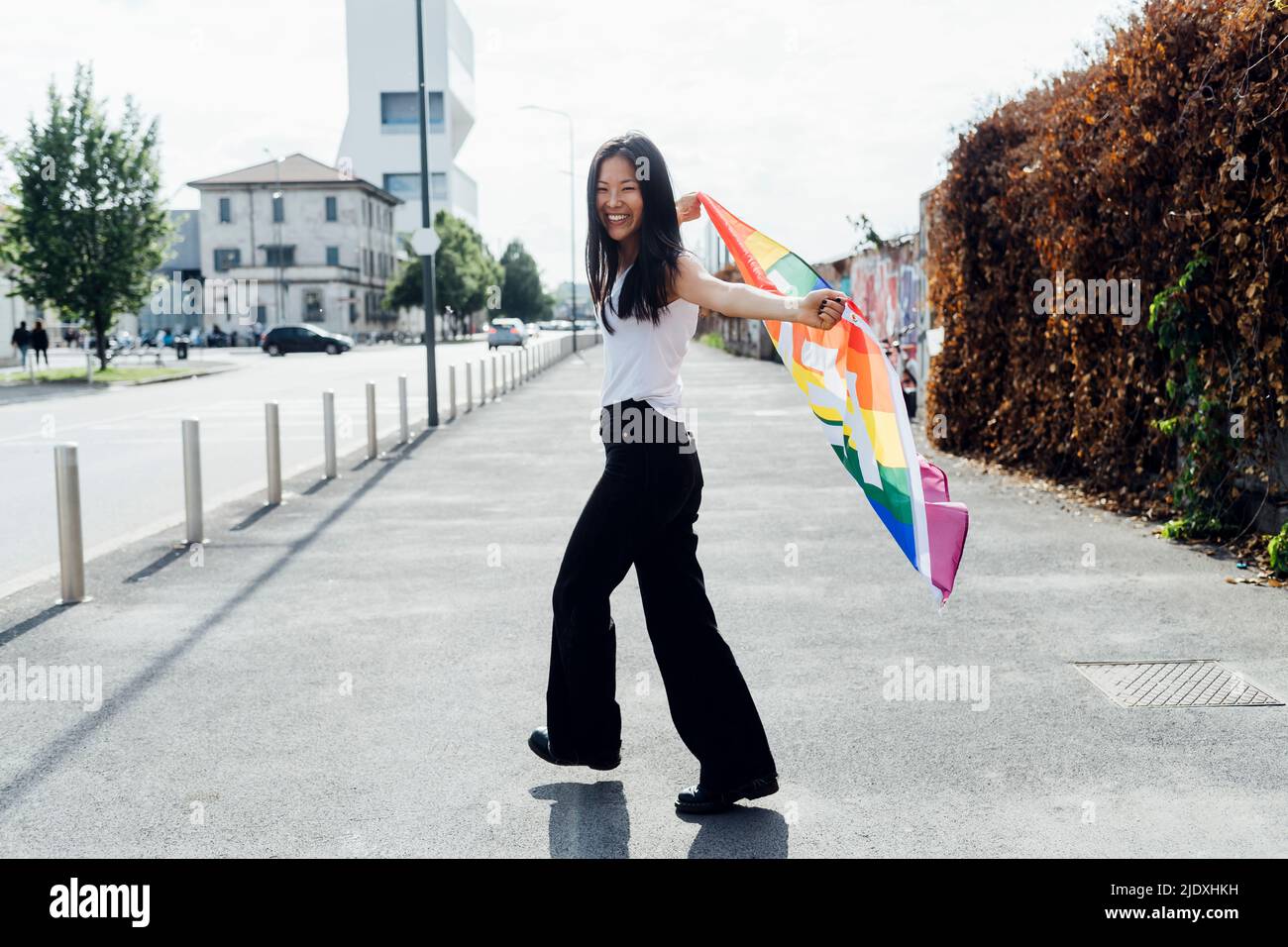 Jeune femme joyeuse avec drapeau arc-en-ciel marchant sur la route Banque D'Images