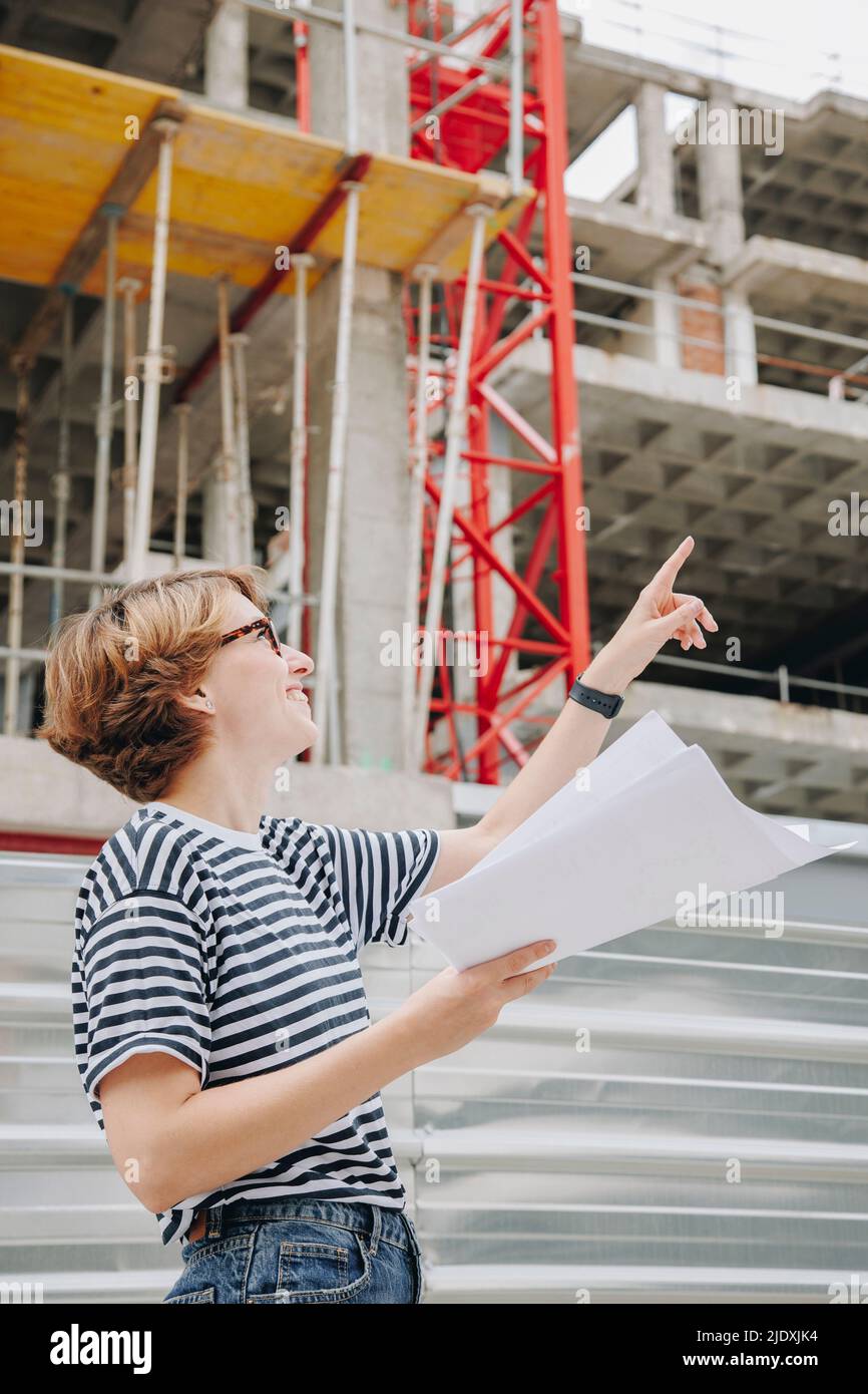 Architecte souriant tenant un plan d'action pointant vers le chantier de construction Banque D'Images