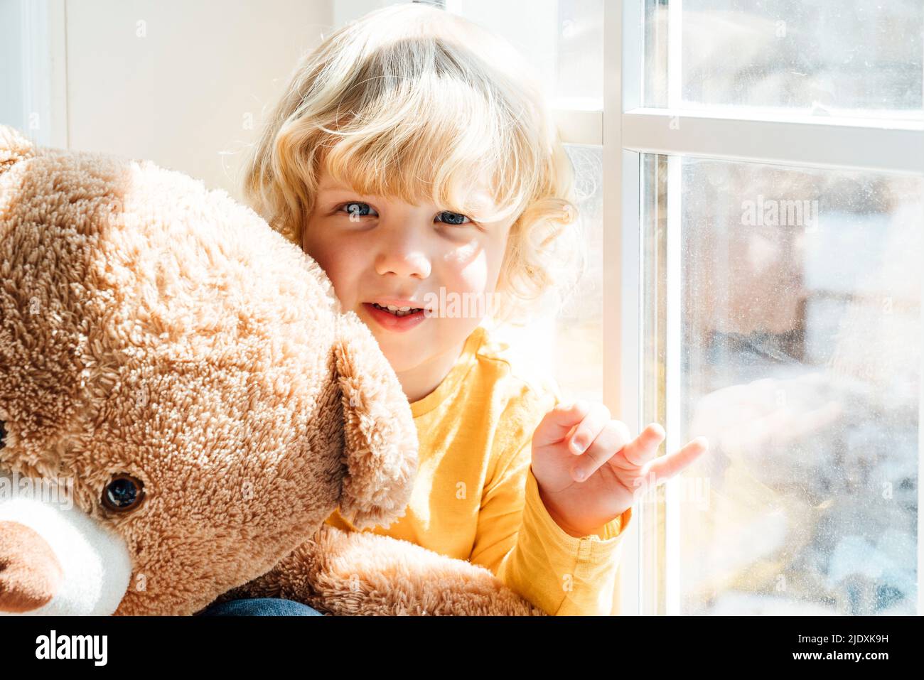 Adorable garçon avec un ours en peluche assis près de la fenêtre à la maison Banque D'Images