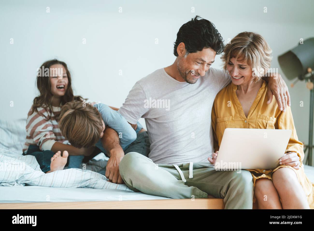 Un homme et une femme heureux avec un ordinateur portable assis par une fille et un fils jouant sur le lit à la maison Banque D'Images