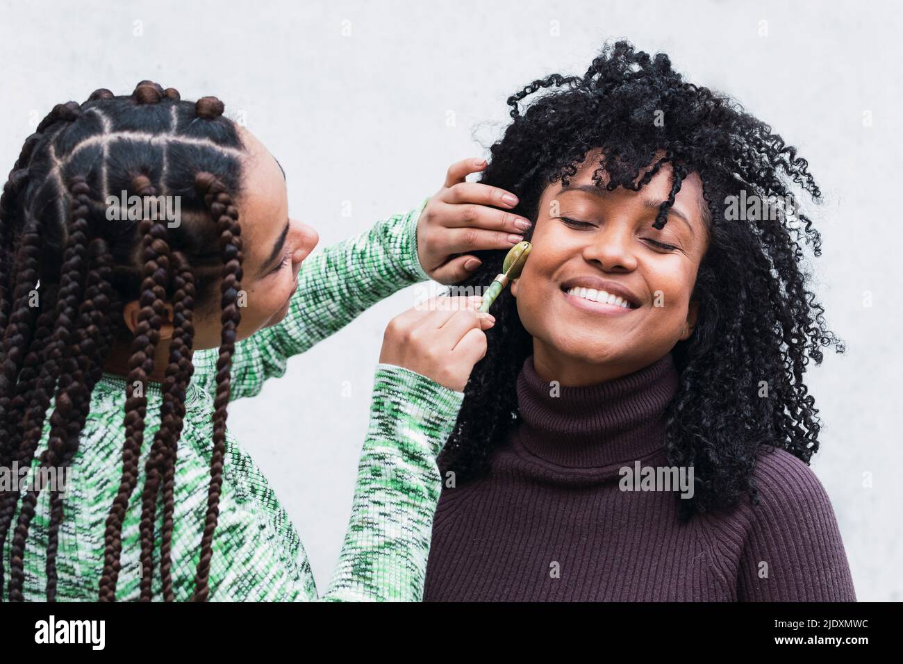 Jeune femme heureuse faisant un massage du visage sur un ami à travers le rouleau de jade sur fond blanc Banque D'Images