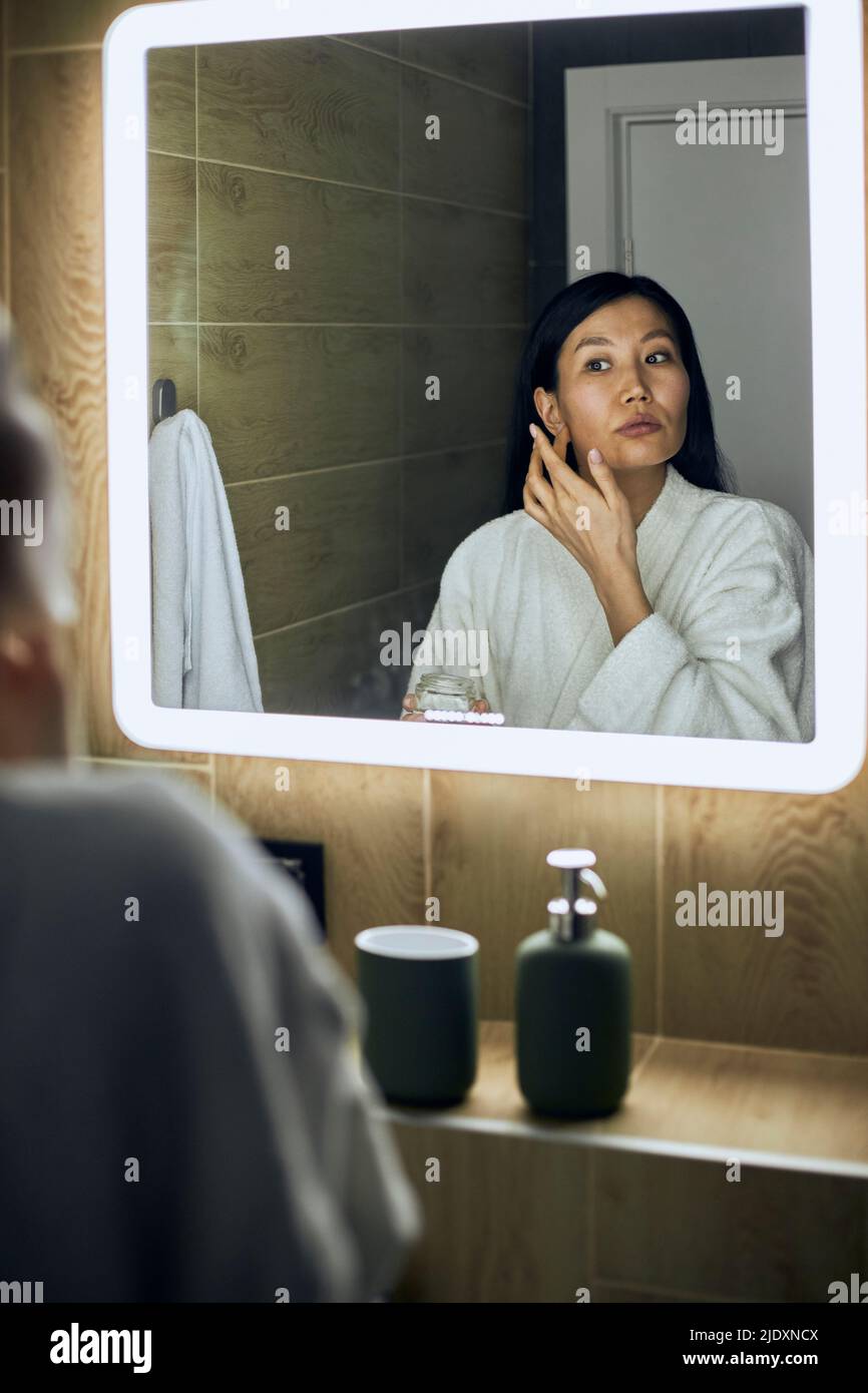 Femme portant un peignoir appliquant de la crème hydratante sur le visage regardant dans le miroir Banque D'Images
