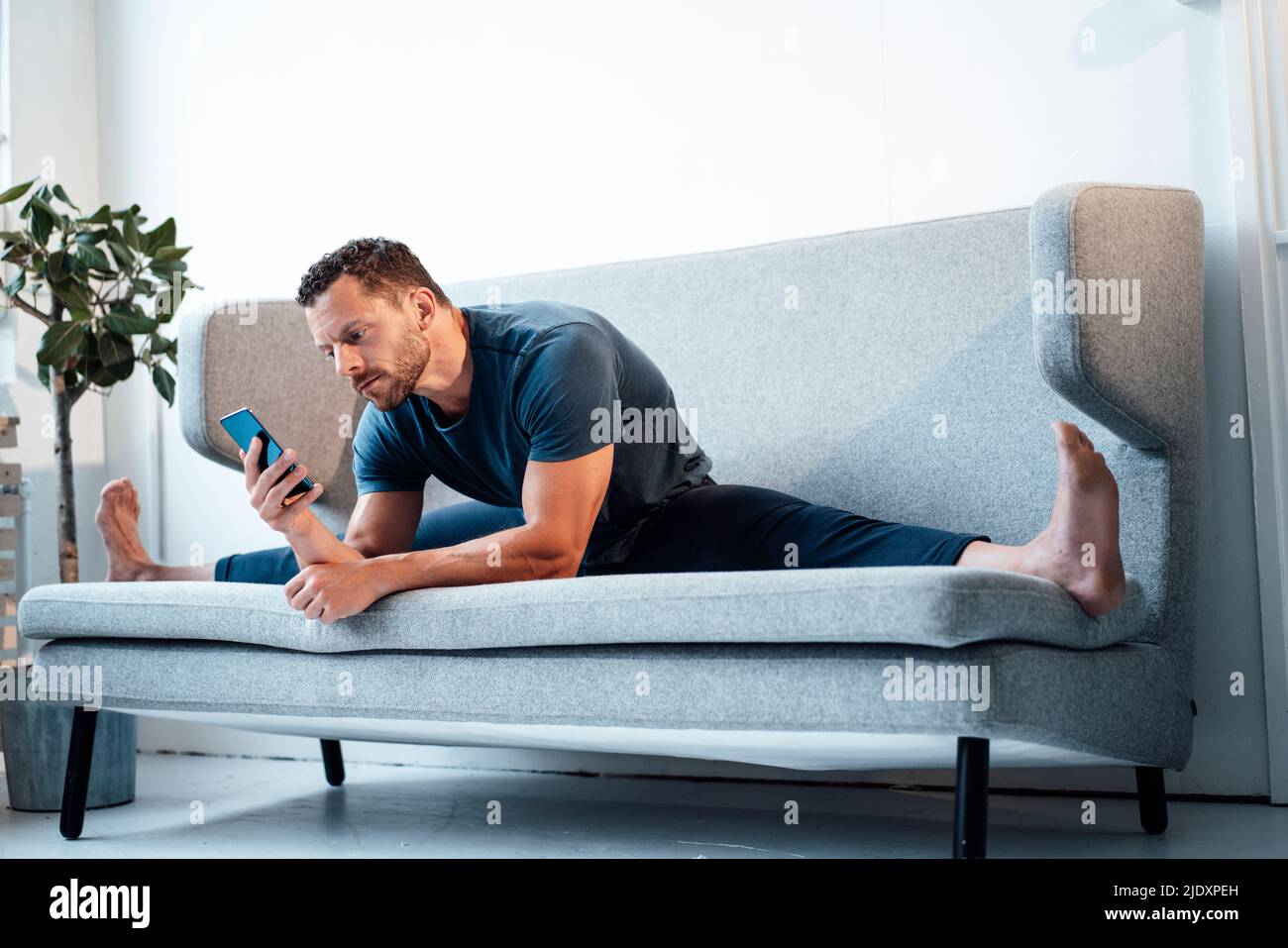 Homme utilisant un smartphone assis avec les jambes séparées sur le canapé à la maison Banque D'Images