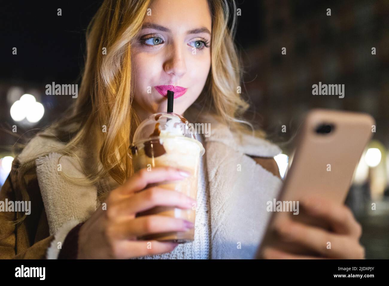 Femme buvant à partir d'une tasse jetable et utilisant un téléphone portable la nuit Banque D'Images