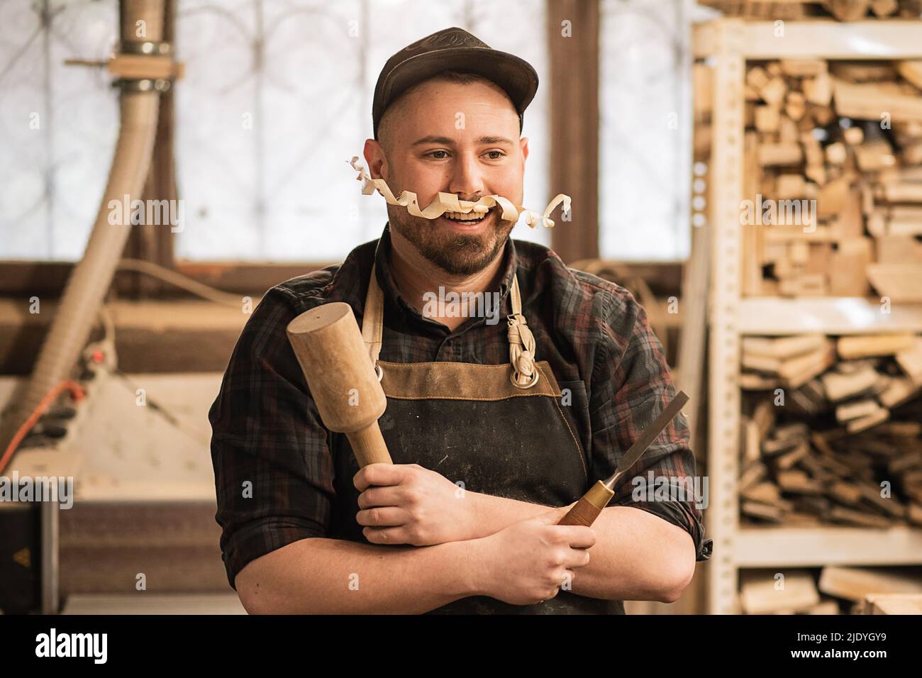 Homme souriant, drôle, menuisier en casquette et combinaison de protection avec moustache en copeaux sous le nez, tenant les outils de construction Banque D'Images