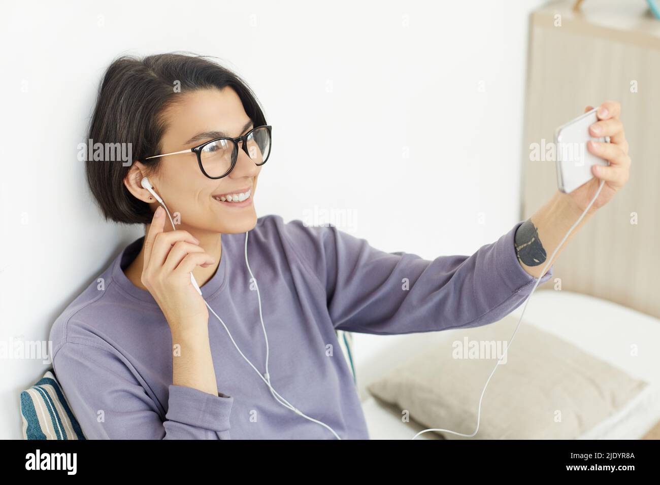 Jolie fille gaie avec tatouage au poignet, avec écouteurs, pour appeler via l'application de visioconférence sur smartphone Banque D'Images
