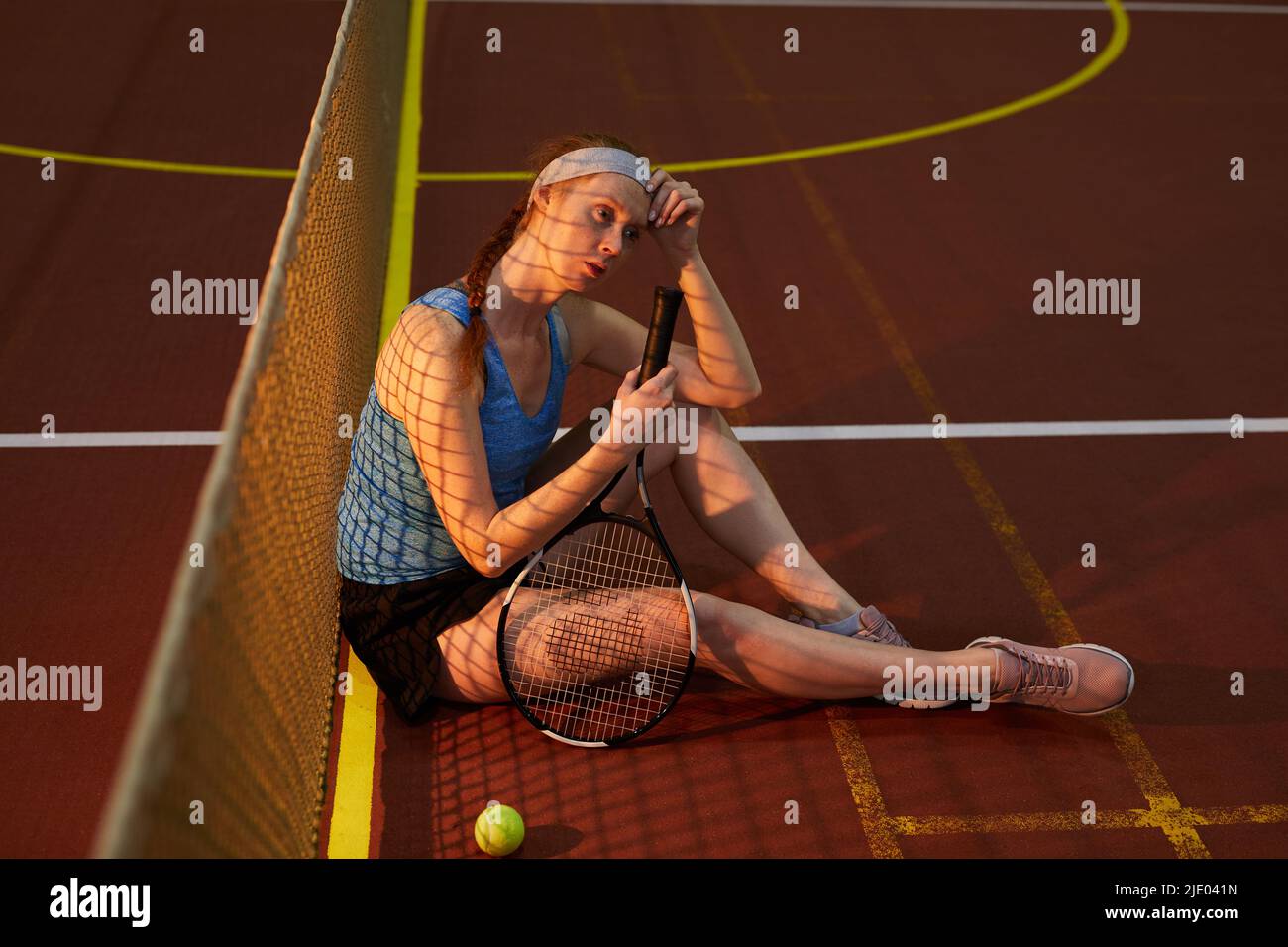 Jeune femme réfléchie à tête rouge avec tresse épuisée après un match de tennis assis sur le sol sur un court de tennis sombre et tenant une raquette Banque D'Images