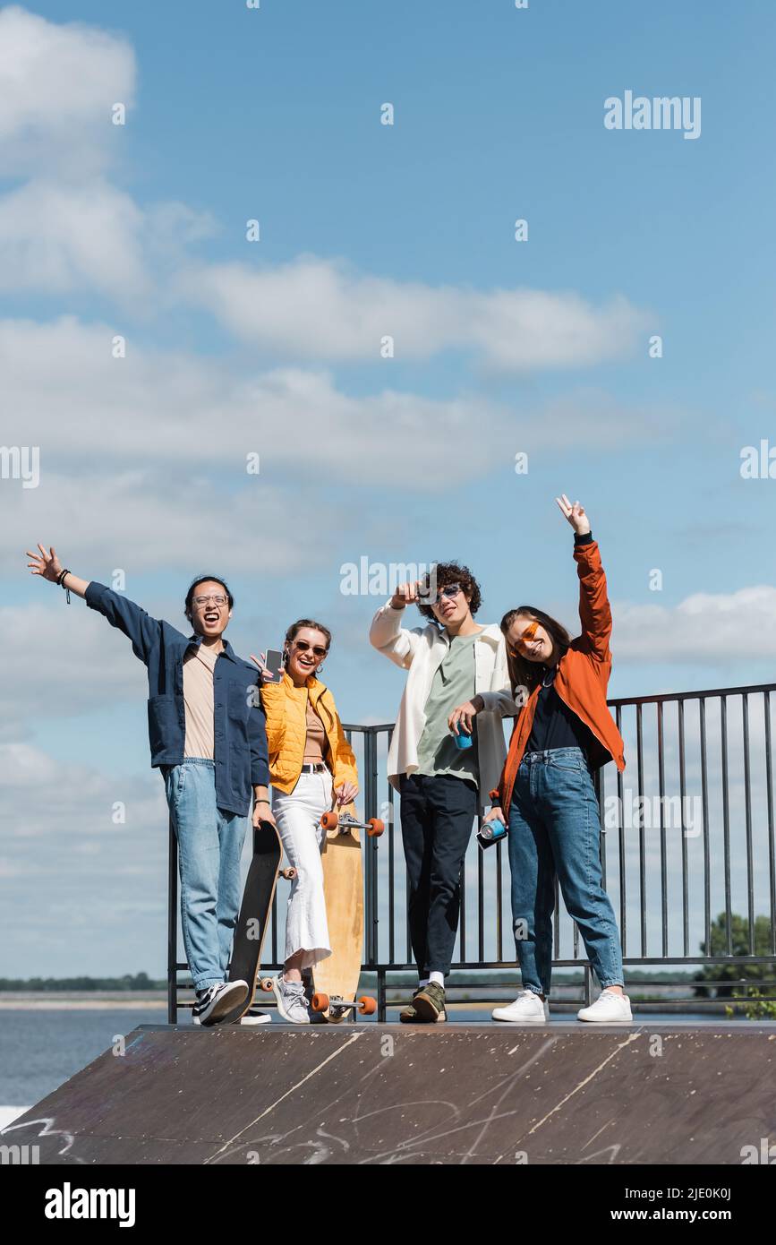 des patineurs multiculturels heureux se sont empatés des mains et ont montré des signes de victoire sur la rampe de skate sous un ciel bleu nuageux Banque D'Images