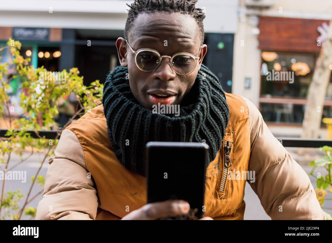 jeune homme africain avec des lunettes, une écharpe noire et des cheveux courts, assis à l'extérieur de la cafétéria, souriant avec des sourcils relevés regardant le téléphone avec Banque D'Images